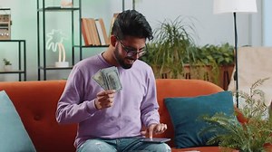 Planning budget. Rich happy indian man counting money cash use smartphone calculator app calculate domestic bills at home. Hindu guy satisfied of income earnings, win, saves money for planned vacation
