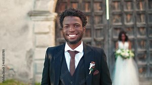 African American man wearing elegant suit with tie positively smiling directly at camera. Attractive bridegroom enjoying happy wedding day. In background standing fiancee with large bouquet of flowers