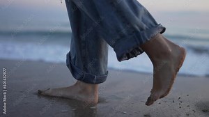 Girl walking barefoot in long jeans along the shore of the beach during sunset. A wave crashes with her feet. Slow motion.