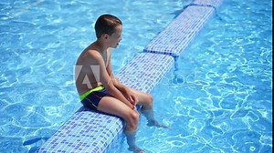 Boy having a rest in the pool. Active boy sitting on the swimming pool after swimming on blue clear water background outdoors. Healthy concept.