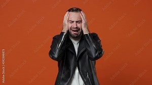 Crying worried young bearded man 20s wearing black leather jacket posing isolated on orange color background in studio. People lifestyle concept. Put palms on head spreading hands ask why screaming