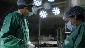 Asian Indian male surgeon or doctor in an green apron is busy operating a surgery using surgical instruments with the help of a female assistant or woman nurse in operation theatre in modern hospital