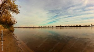 Time lapse on sandy riverbank with fast moving clouds in sky illuminated by orange sunset reflecting in water. Bodies of water, environmental conservation, caring for ecology, limiting harmful