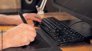 Close up view on graphic designer's hands using tablet and touch screen pen for drawing. One hand clicking buttons on keyboard, another one drawing with pen. Blurred background, soft selective focus