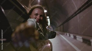 Portrait of a beautiful girl hang out of car window on sunny road trip day. The woman leans out of the car window, stretches her hand, feels the wind with her fingers. Smile of a female in a tunnel