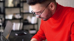Pensive bearded hipster student reading paper reports and writing down information for course work in modern apartment.Smart young man checking data and working remotely at home interior