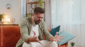 Sad young man feeling hopelessness loneliness, nervous breakdown, crying shouting while sitting on sofa in living room at home. Caucasian guy cries from despair, being sad because of unfair situation.