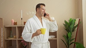 Middle-aged man in a spa room wearing a white robe holding a yellow cup while talking on the phone surrounded by candles and plants