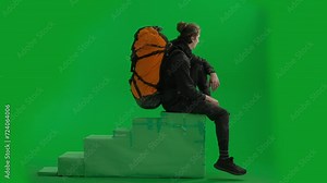 Side view of a male traveler with a backpack on his back, sitting on an elevated position on a green screen. The man sits with his legs overhanging, puts his hand to his eyes, stares into the distance