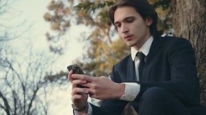 Confident man in a business suit is sitting outdoors and using smartphone. A young man in a suit with mobile phone, slow motion. Serious and calm businessman in the city looking to a cell phone.