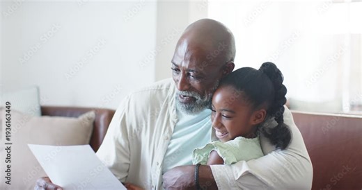 African American senior man reading paper while child giggling and hugging, sharing news on sofa