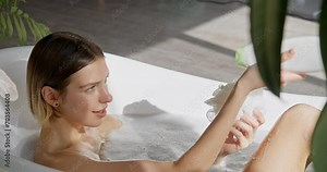 Young blonde woman pouring body wash on sponge, while sitting in bathtub. Side view of seductive female scrubbing legs with sponge, while taking bath at home. Self care, hygiene, cosmetics concept.