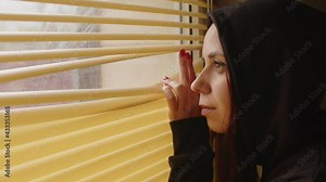 Close up of young woman watching through window blinds at home. Portrait of curious female observing through window jalousie in room.
