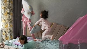 Teen girls have pillow fight three little sisters asian girls jumping on bed together. Slow motion.