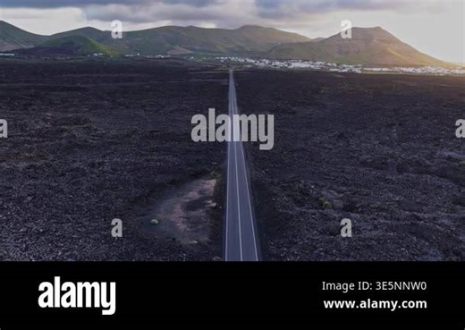 A road stretches through a volcanic landscape in Lanzarote, Spain. Fields and mountains surround the path. A small town is visible in the distance under cloudy skies around sunset Stock Video Footage - Alamy