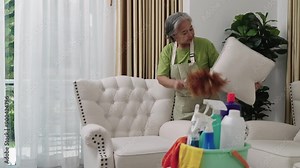 Handheld Shot Footage shows a housekeeper wearing an apron dusting and arranging cushions on a sofa in a pristine living room. Her dedication guarantees a clean and organized home.