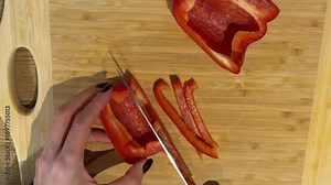 Slicing a Red Bell Pepper into Strips on a Wooden Board. A top-down view shows hands slicing a red bell pepper into thin strips on a wooden board, preparing fresh vegetables with a sharp knife.