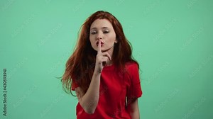 Smiling woman with red hair holding finger on her lips over green background. Gesture of shhh, secret, silence. Close up