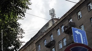 The wind shakes a blue road sign with a left direction arrow against the backdrop of city buildings and residential high-rise buildings. City atmosphere. Telephone antenna on a radio tower