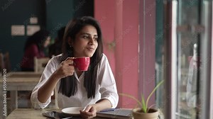 young Asian woman siting holding coffee on chair in the coffee shop.Thoughtful young ethnic Indian woman make plan or decide working with looking the windows.