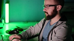 Programmer guy with a beard in glasses works at a computer in a room against the background of a green lamp. Side view