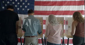 Four voters casting ballots at election booths, seen from behind. Wide shot with faces mostly hidden by camera angle. United States flag on wall in background. 4K DCI Stock Video