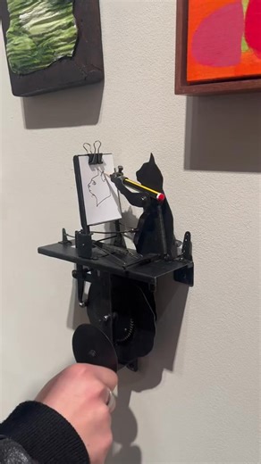 The charming kinetic artwork called Shadow of the Cat is created by British artist Tim Lewis. It features a whimsical cat-shaped figure that mechanically draws the outline of a cat's head (or portrait) on paper using a pencil, powered by springs, gears, and clever engineering. This piece is inspired by Jean Tinguely's iconic Méta-matic series from 1959—self-operating drawing machines made from scrap metal, motors, and random mechanisms that produced abstract marks on paper. | GIGadgets