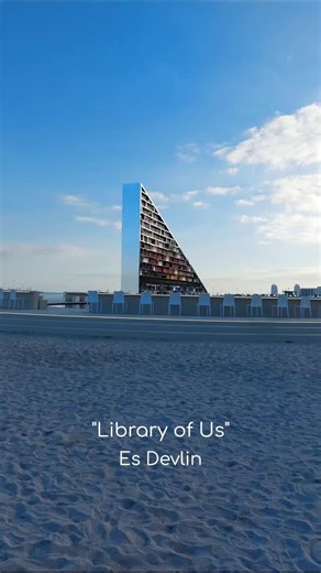 Women in the Arts Inc. on Instagram: "Maria Guerrero shares a quiet moment with Es Devlin's installation "Library of Us" and the imprints of a community. 'The sunlight is dancing away as my feet sink into the warm and timid-yellow sand. From the distance, and between me and the overlapping flat, horizontal brushstrokes of sea and sky, the triangular shaped library rises powerful above water, yet delicately balanced, motionless on its circular base, three steps above the ground . . .' Read the fu