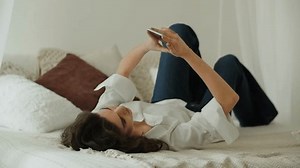 Brunette girl lying on her bed and using smartphone. Young woman texting message in the bedroom