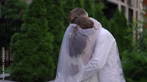A bride kisses her groom while covering him with her veil. A couple of newlyweds kissing under veils, standing outdoors on a summer day against the background of green trees on the territory of a