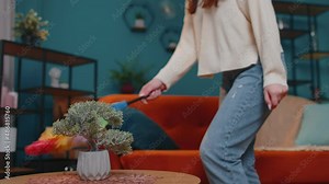Joyful young woman dusting furniture caring for hygiene using colorful duster in living room having fun listening to music on headphones, dancing. Girl singing into duster like into microphone at home