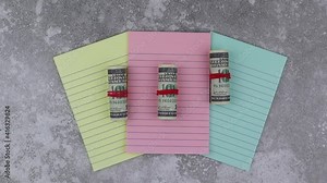 Rolls of 100 dollar banknotes on colored notepads arranged fanned to the centre of the screen on a wood background in an overhead view for financial concepts. Stop motion, top view