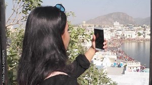 Handheld woman takes selfie photo video with her mobile cellphone device from a panoramic vantage view point of holy Pushkar lake, a sacred city for Hindu pilgrims mela festival bathing ghats temples