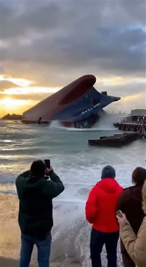 A gigantic ship is rapidly sinking near the shore while small boats rush around and people on beach
