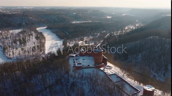 Aerial daylight sweep of Turaida Castle in Sigulda, Latvia, its red brick towers and walls dusted with snow as a drone tracks over the frozen Gauja River.