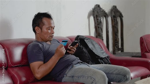 Asian man at home sitting in sofa. Male client banking buying shopping online with credit bank card and mobile smartphone, e-commerce order delivery payment internet pay.