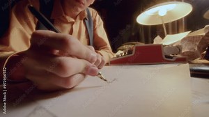 Hands of young man writing letter on sheet of paper with fountain pen at desk with warmly lit lamp and vintage typewriter. Close-up view
