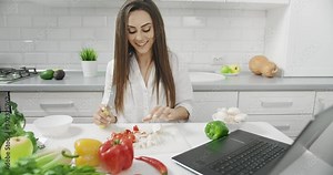 Beautiful young female blogger slicing vegetables in front of laptop camera, recording video for channel, vegan lifestyle