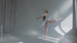 Female dancer does ballet exercises in stage dress with open back. Rehearses dance moves in the spacious studio bathed in sunlight. Slow motion.