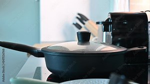 Large frying pan with a glass lid, steam from cooking. A woman opens a pot and stirs food. The process of making homemade lunch. Vegetables are fried. Eggplant Vegetarian Lunch