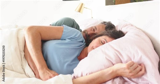 Senior couple spooning in bedroom, woman appearing asleep on pink pillow, man blinking and settling