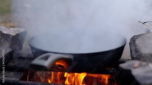 Steaming Skillet on a Blazing Bonfire in the Woods. Flaming, burnt out, frying pan in a fire, standing on a metal grate. Smoke, soot, fog over a bright flame evening. Outdoor cooking. Camping. Hiking.