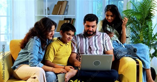 Indian young family using laptop computer at home sitting together on sofa while surfing internet and shopping online indoors inside a modern lavish living room enjoying digital lifestyle