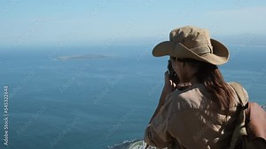 traveler in safari clothes and a hat takes a photo of the ocean from a high mountain. a hiker in boots and shorts climbed a mountain pass and takes a photo of the mountains. photographer with camera