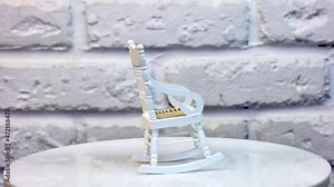 Little white rocking chair for doll’s house. Pretty small piece of furniture on the rotating table. White brickwall at the background. Close up.