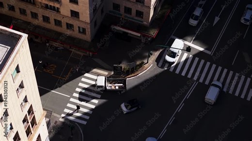 Top-down drone view in Genoa, Liguria, Italy, showing vehicles turning at an intersection as a truck stops to let a pedestrian cross under strong light and shadow contrast.