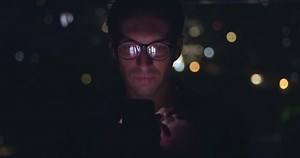 Portrait of young man using mobile phone, scrolling app pages social networks, mobile screen reflected on his face. Male in glasses scrolling internet on mobile phone background of night city.