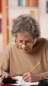 Vertical video of frustrated angry upset elderly senior old woman using pencil doing paperwork and laptop, doing taxes, unpaid bills, huge taxes and high electricity rates, insufficient pension.