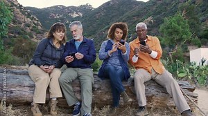 Group of senior friends on hike in countryside sitting on log checking mobiles phones for fear of missing out - shot in slow motion