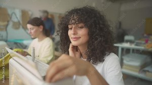 Side view of pretty Caucasian woman with curly hair sitting in front of sewing machine. Placing spool of thread on top of machine and adjusting everything before starting working in morning.